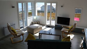 A living room with a sofa, two armchairs, a coffee table, and a TV. There are sliding glass doors leading to a balcony with a view of houses and a beach. - Le Perchoir Avec vue Mer, à 300m de la Plage (Saint-Côme-de-Fresné)
