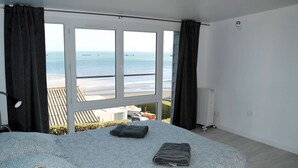 A bedroom with a large window overlooking a beach, a bed with a patterned bedspread, and a radiator on the wall. - Le Perchoir Avec vue Mer, à 300m de la Plage (Saint-Côme-de-Fresné)