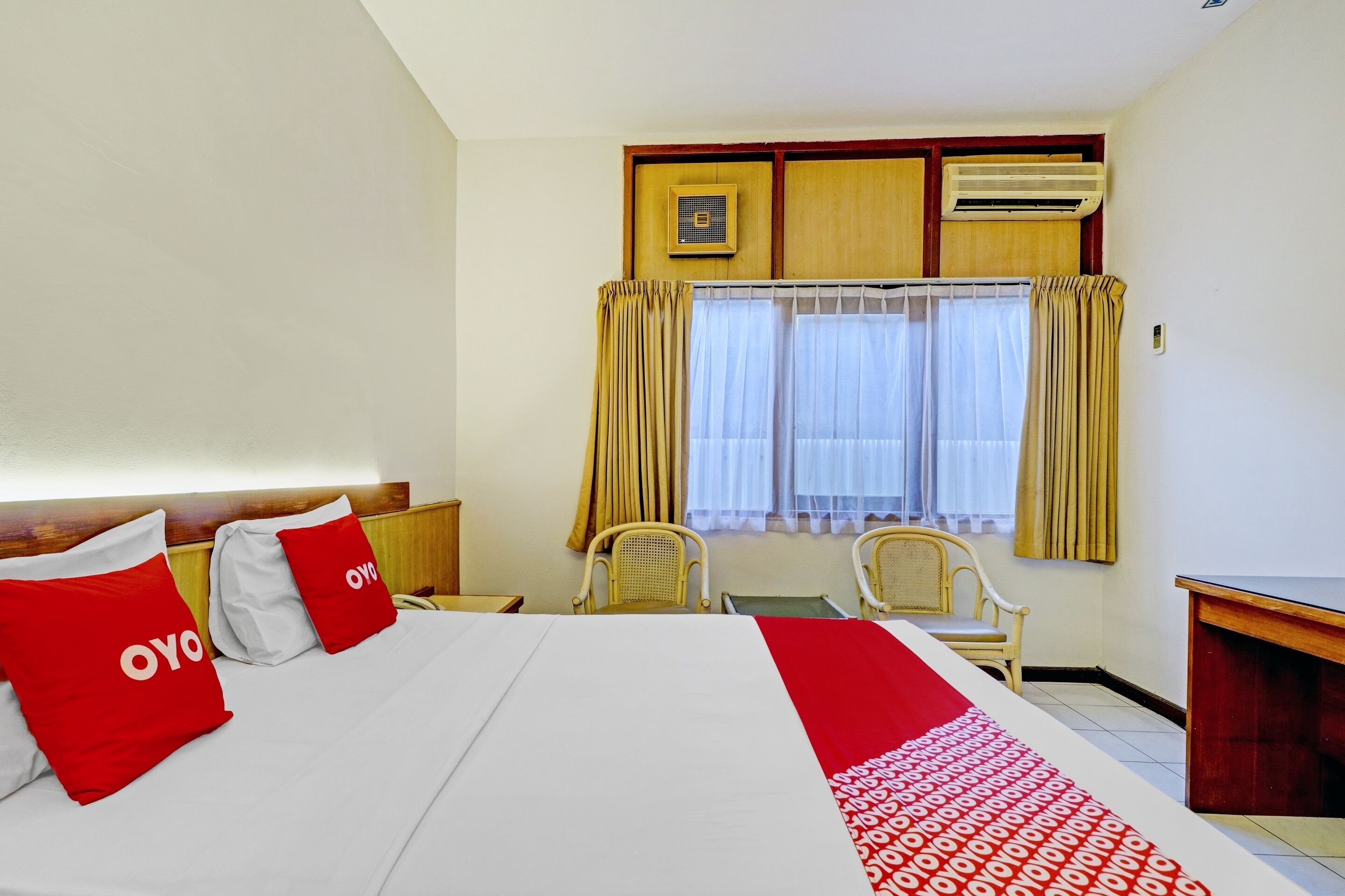 A hotel room with a large bed, two red pillows with "OYO" branding, a wooden headboard, a desk, two chairs, and a window with curtains.