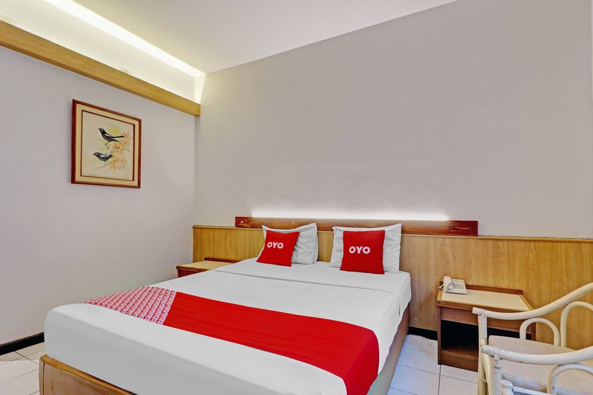 Habitación de hotel con una cama, dos almohadas rojas, cabecera de madera, mesita de noche con un teléfono, una silla e imagen enmarcada en la pared.