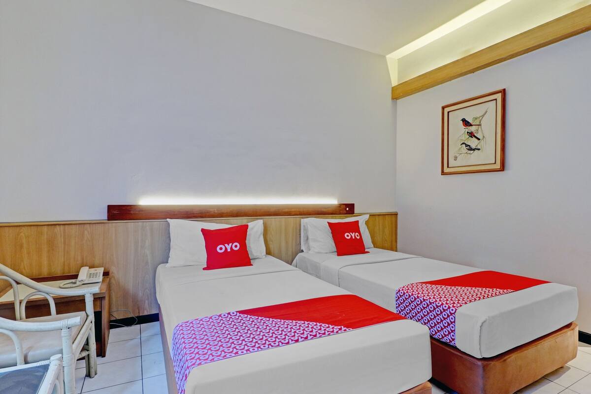 Habitación de hotel con dos camas, cada una con un cojín rojo con la etiqueta "OYO", un cabecero de madera, una mesita de noche con un teléfono y un cuadro en la pared.