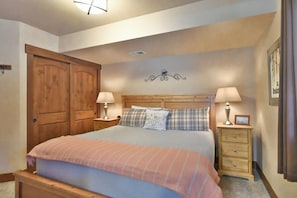 A bedroom with a large bed, two bedside tables with lamps, a wooden wardrobe, and a wall decoration.