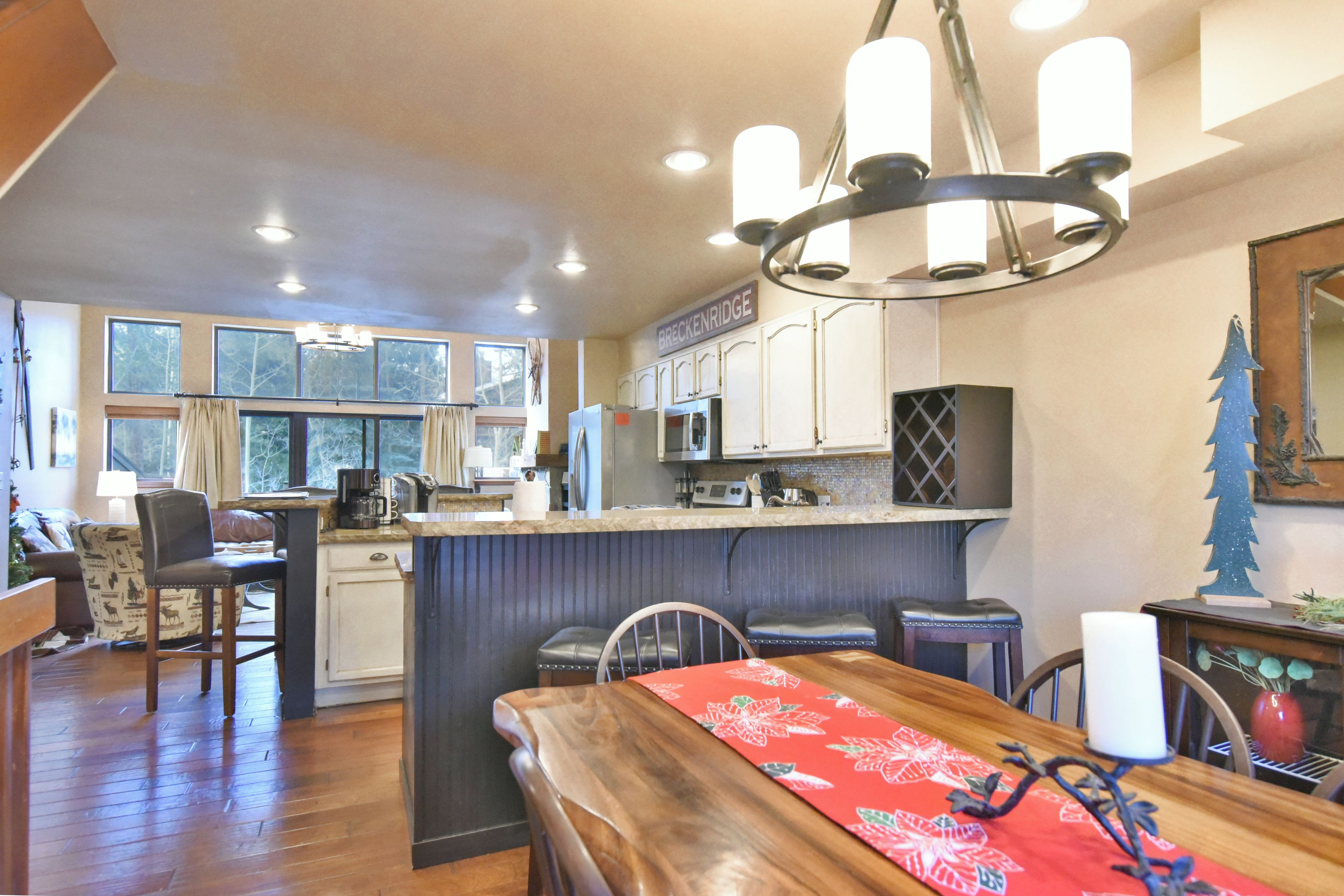 A kitchen with a central island, a dining area with a wooden table, and a view of the outdoors through large windows.