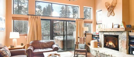 A cozy living room with a fireplace, leather sofa, and a large window overlooking a forest.