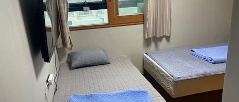 Une petite pièce bien éclairée, avec deux lits : l’un est recouvert d’une couette bleue et l’autre d’une couette grise. Il y a une fenêtre avec des rideaux, un téléviseur fixé au mur et une prise de courant sur le côté gauche.