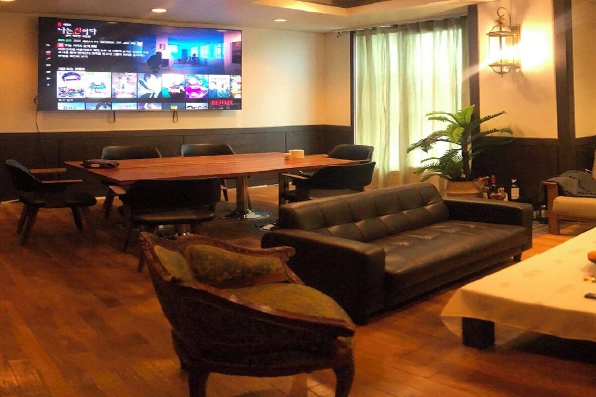 Ein Hotelzimmer mit einem Holztisch, einer Ledersofa und einem an der Wand befestigten Fernseher, der mehrere Sender zeigt.