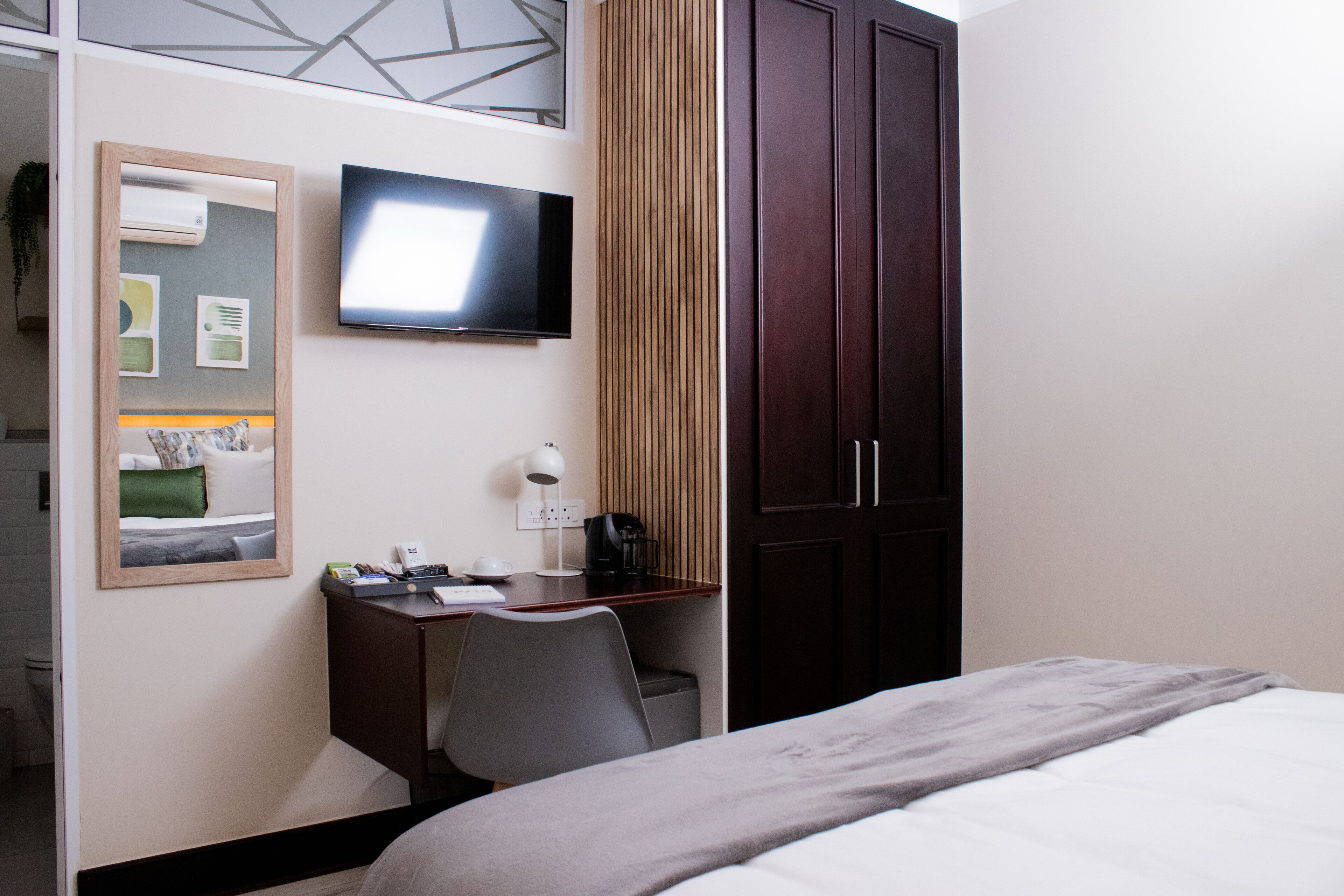 A hotel room with a bed, a desk with a chair, a television, a mirror, and a wardrobe.