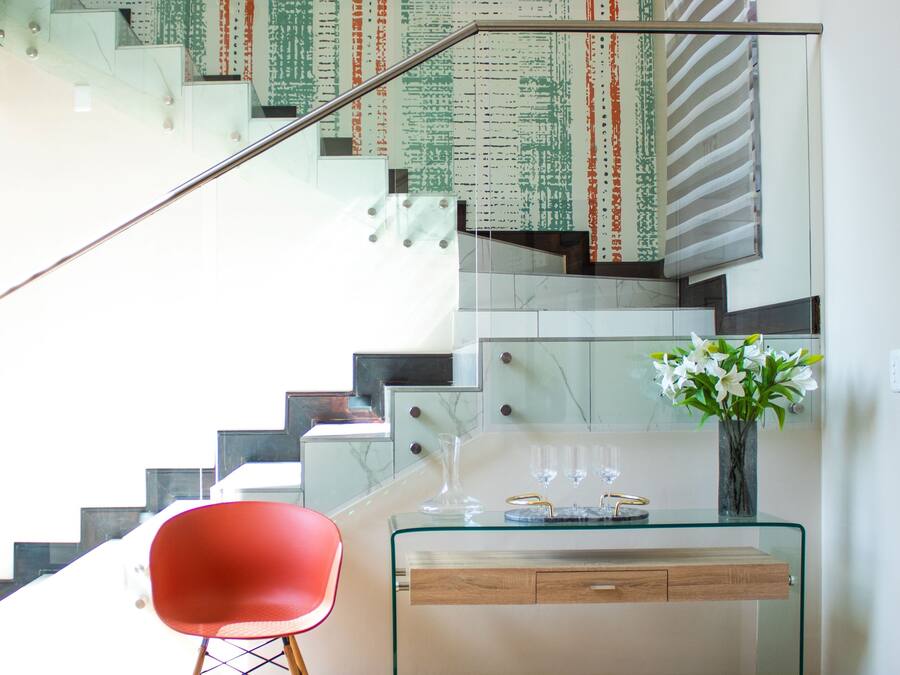 Un interior moderno con un escritorio de vidrio, una silla roja, una escalera y un jarrón con flores.