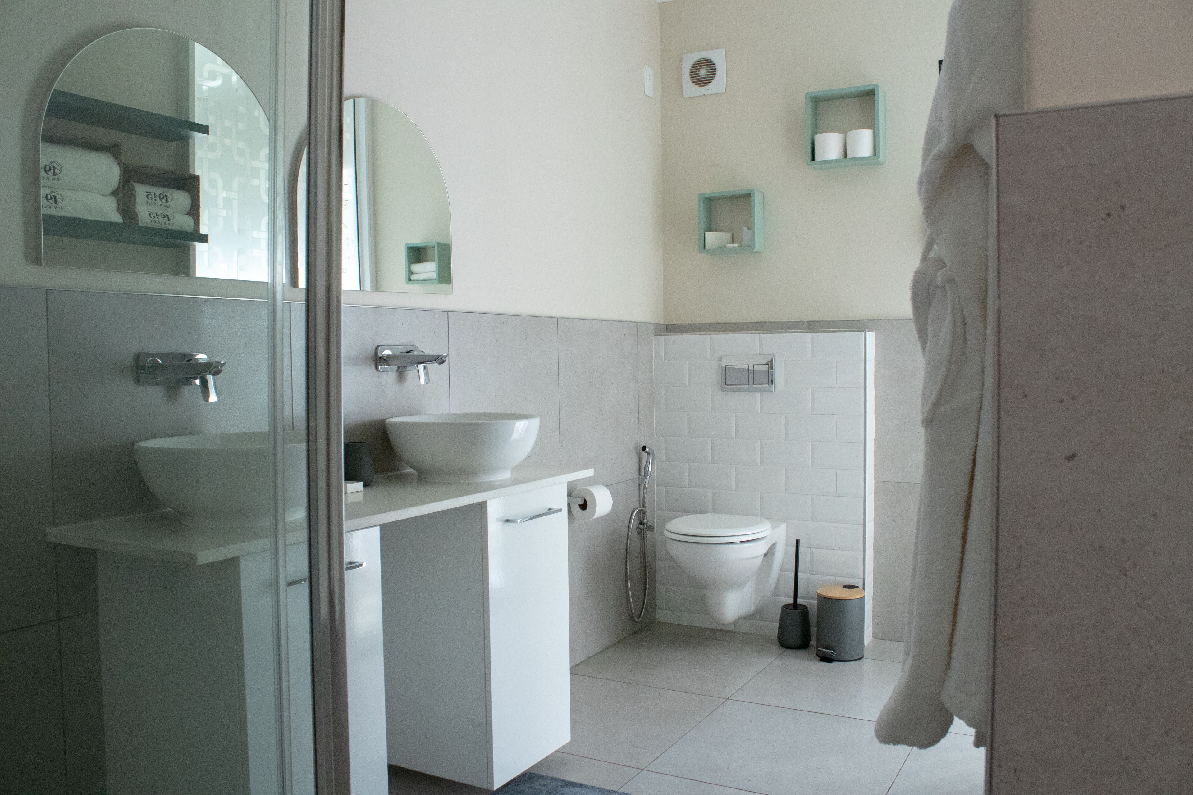 Un baño moderno con un lavamanos, inodoro y ducha blancos. Hay repisas con artículos de aseo personal y una toalla colgada en la pared.