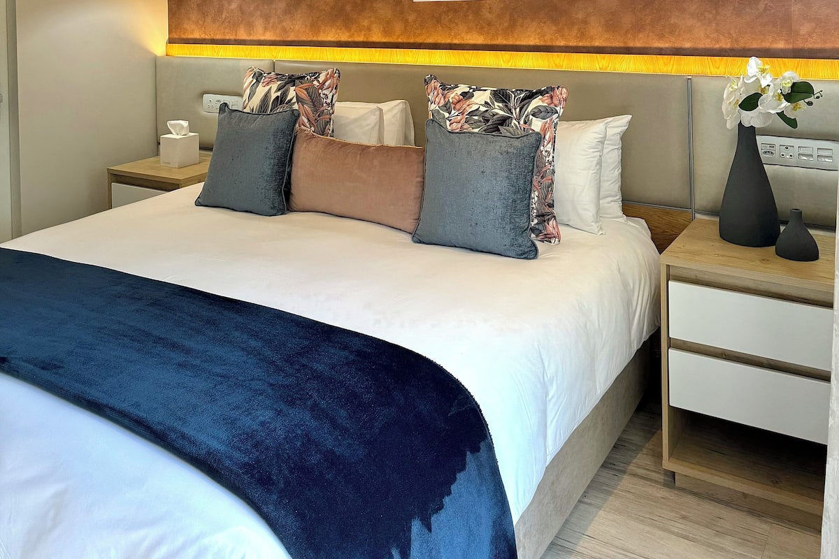 Une chambre d’hôtel moderne avec un grand lit, deux tables de chevet, une petite table, un appareil de climatisation et des œuvres d’art accrochées au mur.
