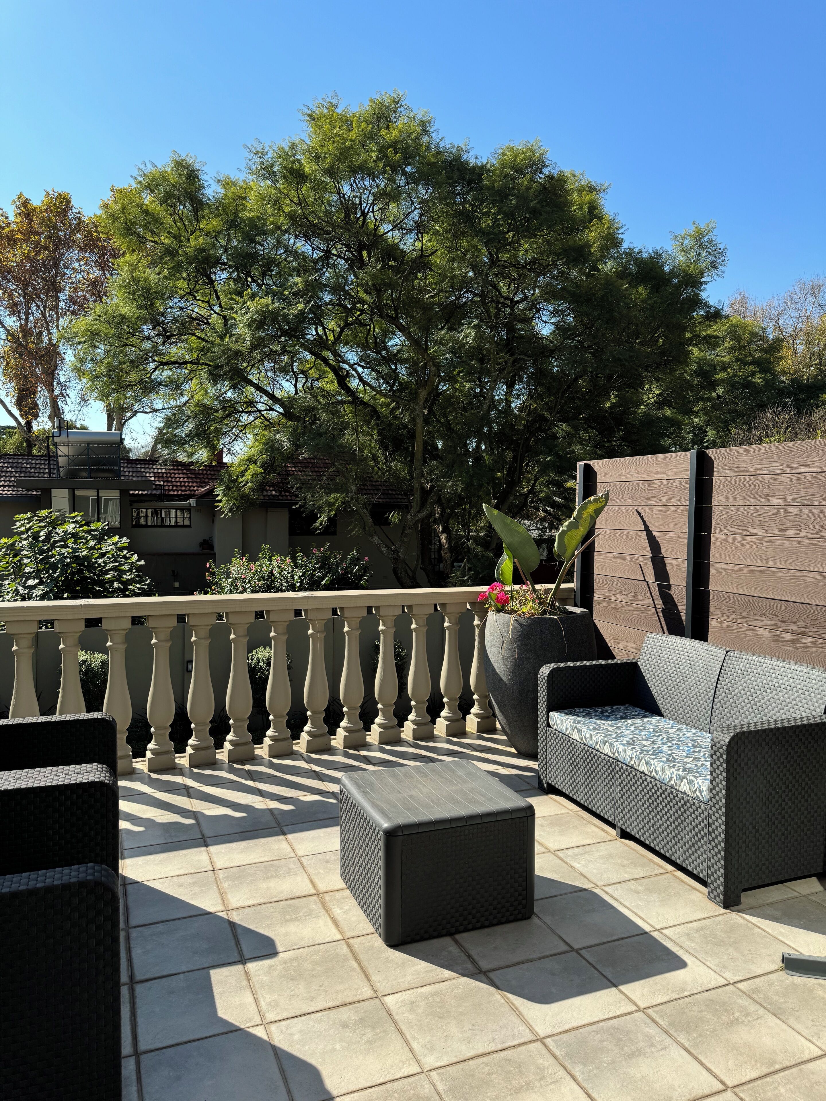 Une terrasse extérieure avec des meubles en osier, un sol carrelé et une vue sur des arbres et un bâtiment en arrière-plan.