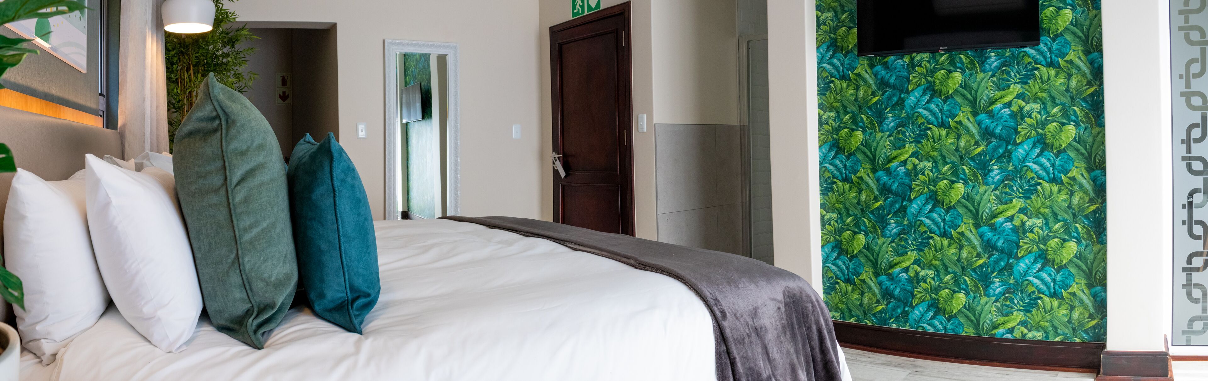 A hotel room with a bed, pillows, a TV, and a wall with a tropical leaf pattern.