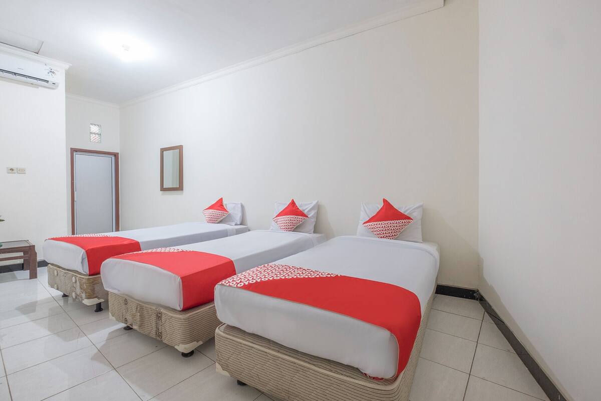 A hotel room with three single beds, each with a red and white bedspread and matching pillows. The room has white walls, a tiled floor, and a wooden door.