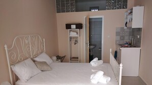 A neatly made bed with white linens and a metal headboard, a small bedside table with a lamp, and a white dresser with drawers. -  Aphrodites Apartments (Syros)