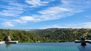 Ein klarer, blauer Himmel mit Wolken, ein Schwimmbecken mit klarem Wasser und Blick auf einen bewaldeten Hügel.