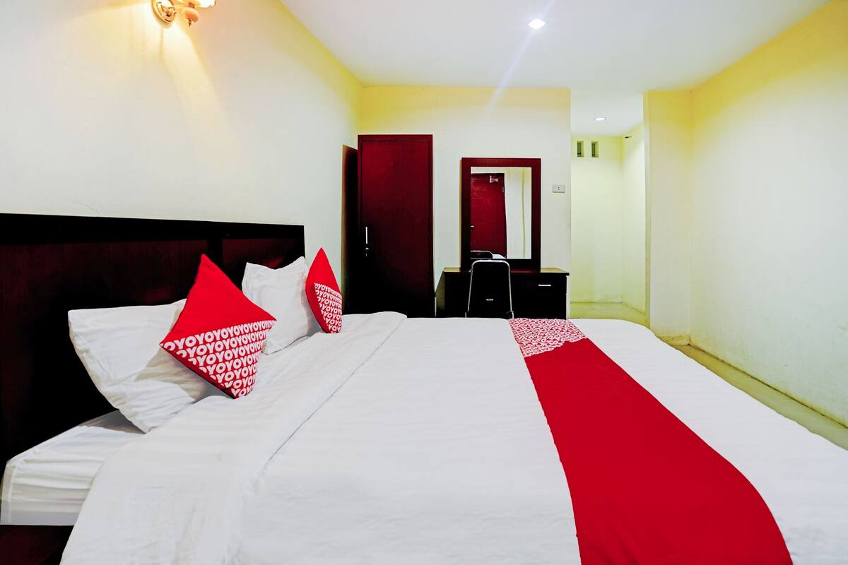 Ein Hotelzimmer mit einem großen Bett, roten und weißen Kissen, einem Holzkopfteil, einem Schreibtisch mit Stuhl, einem Spiegel und einem Kleiderschrank.