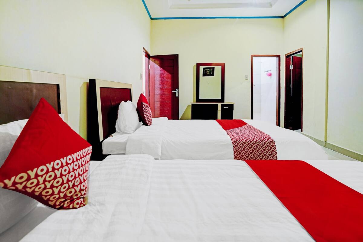 Ein Hotelzimmer mit drei Betten, jedes mit weißer Bettwäsche und roten Akzenten. Auf dem Bett rechts liegt ein Bettläufer mit rotem und weißem Muster. Im Zimmer befindet sich ein kleiner Schreibtisch mit einem Spiegel darüber, und im Hintergrund ist eine Tür mit rotem Rahmen zu sehen.