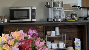 Eine Küchenzeile mit Mikrowelle, Kaffeemaschine und Kaffeekanne. Ein Fenster mit Jalousien, ein Ablagefach mit Kaffeetassen und ein Blumenstrauß.