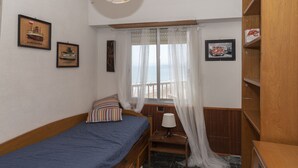 A bedroom with a single bed, a nightstand, a lamp, a window with sheer curtains, and a view of the sea. - EL RACO, A-8º-16 (Cullera)