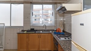 A kitchen with wooden cabinets, a granite countertop, a stove, a sink, and a window with a view of buildings. - EL RACO, A-8º-16 (Cullera)