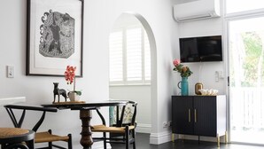 Un coin repas avec une table ronde, des chaises, un parquet en bois foncé, un tapis à motifs, une œuvre d’art encadrée, un vase avec des fleurs et un téléviseur fixé au mur.
