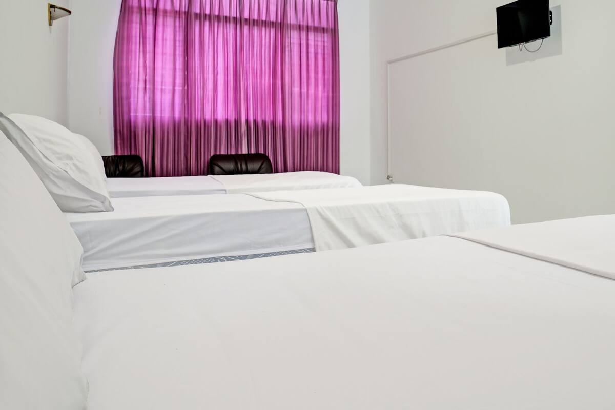 Een hotelkamer met twee eenpersoonsbedden, een zwarte leren fauteuil, een aan de muur bevestigde televisie en een raam met roze gordijnen.