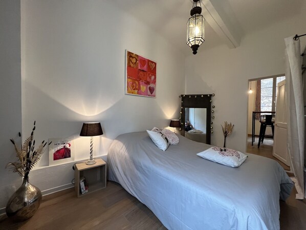 A bedroom with a large bed, a bedside table with a lamp, a mirror with lights, and a window with curtains.