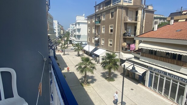 A balcony with a white plastic chair, a view of a street with buildings, palm trees, and a shop named 'Foto Loren'. - B&B HOTEL LUCCIOLA (Cattolica)