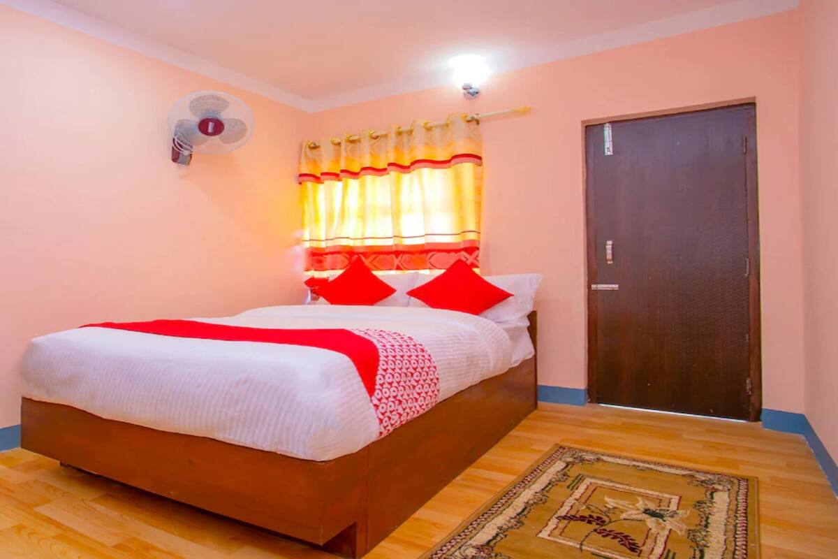 Un dormitorio con una cama de madera, una colcha roja y blanca, un ventilador de pared y una alfombra estampada.