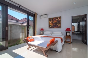 A bedroom with a bed, a wooden chair, a nightstand, a painting, and a bathroom visible through an open door. - Villa Anna (Banjar)