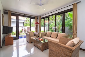 A spacious living room with a sofa, two armchairs, a coffee table, and a TV. Large windows offer views of a pool and greenery. - Villa Anna (Banjar)