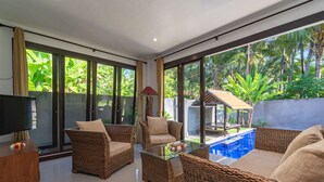 Ampla sala de estar com móveis de vime, mesa de centro de vidro e vista para uma piscina rodeada por vegetação exuberante.