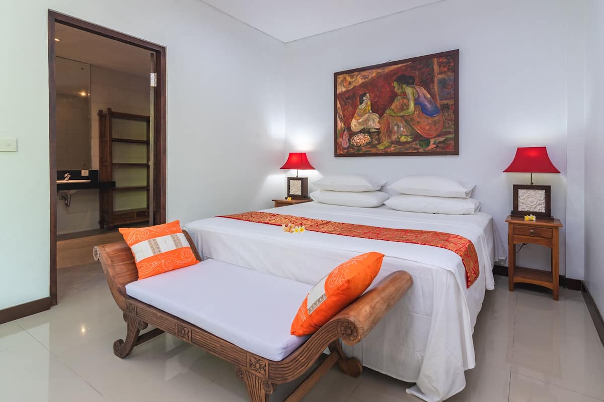 Un dormitorio con una cama, dos mesitas de noche, un banco de madera, un cuadro y una lámpara roja.
