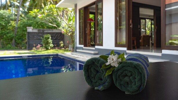 Vista da piscina com uma toalha e uma flor sobre uma mesa, uma casa com grandes janelas e um jardim exuberante.
