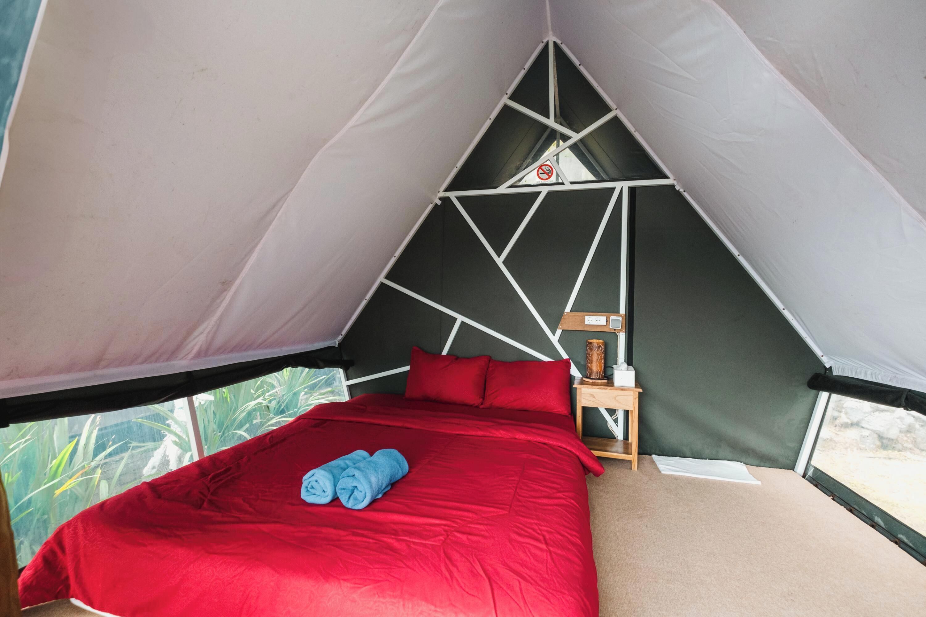 A tented accommodation with a bed, red bedding, and a small bedside table.