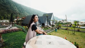 Une femme est assise à une table, devant laquelle s’étend un paysage montagneux, avec une étendue d’eau en arrière-plan.