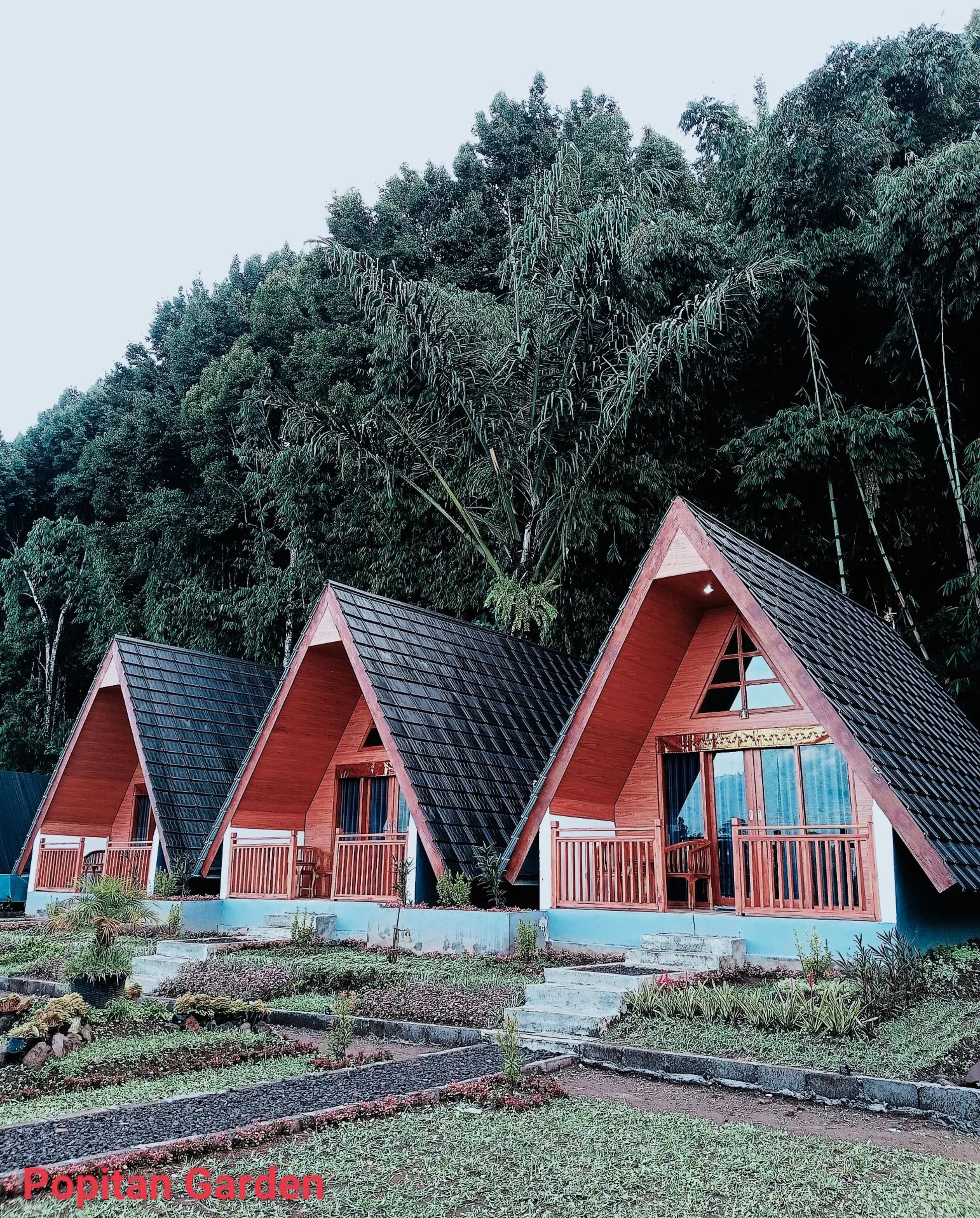 Três cabanas com telhados triangulares e guarda-corpos de madeira, situadas num jardim exuberante com uma floresta densa ao fundo.