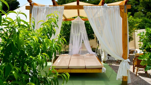 A wooden canopy bed with white curtains and a green mat, surrounded by lush greenery.