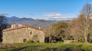 A stone building with a sloped roof, surrounded by trees and mountains in the background.