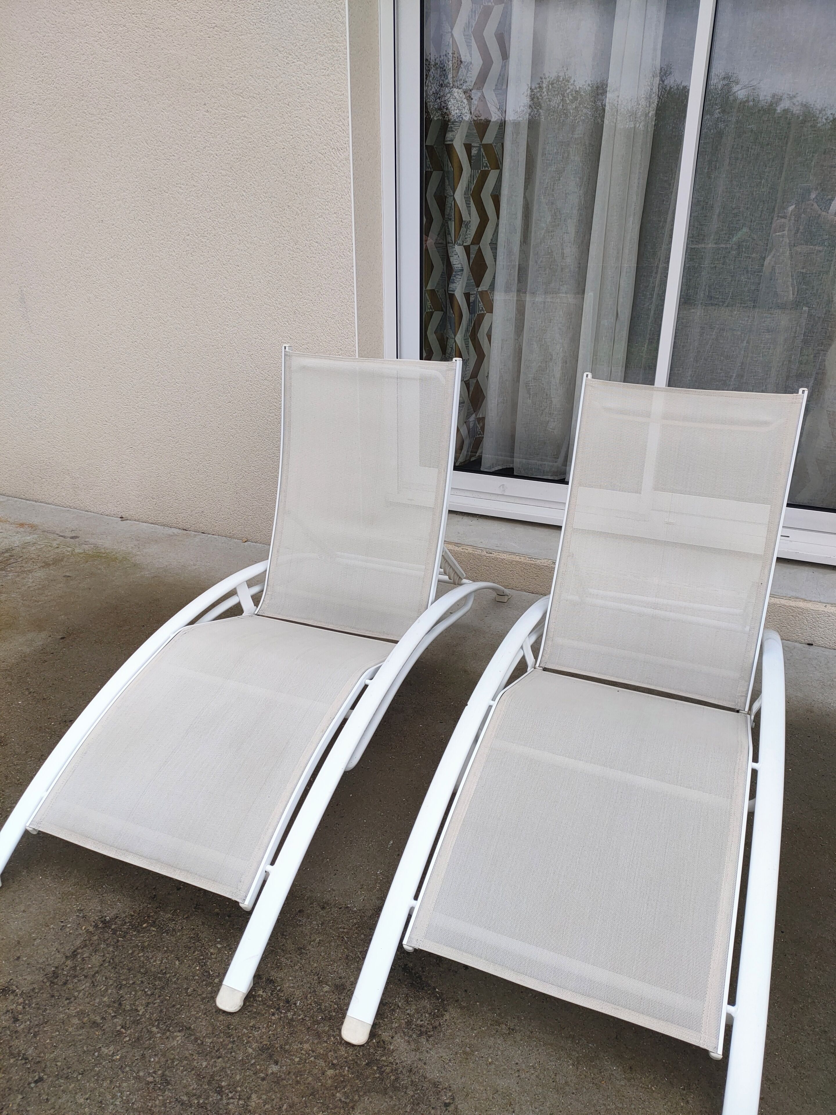 Zwei weiße Lounge-Stühle mit Mesh-Rückenlehnen und -Sitzflächen stehen auf einer Beton-Terrasse neben einem Gebäude mit einem Fenster und einem gemusterten Vorhang.
