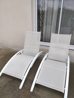 Dos sillones blancos con respaldo y asiento de malla, ubicados en un patio de concreto al lado de un edificio con una ventana y cortina estampada.