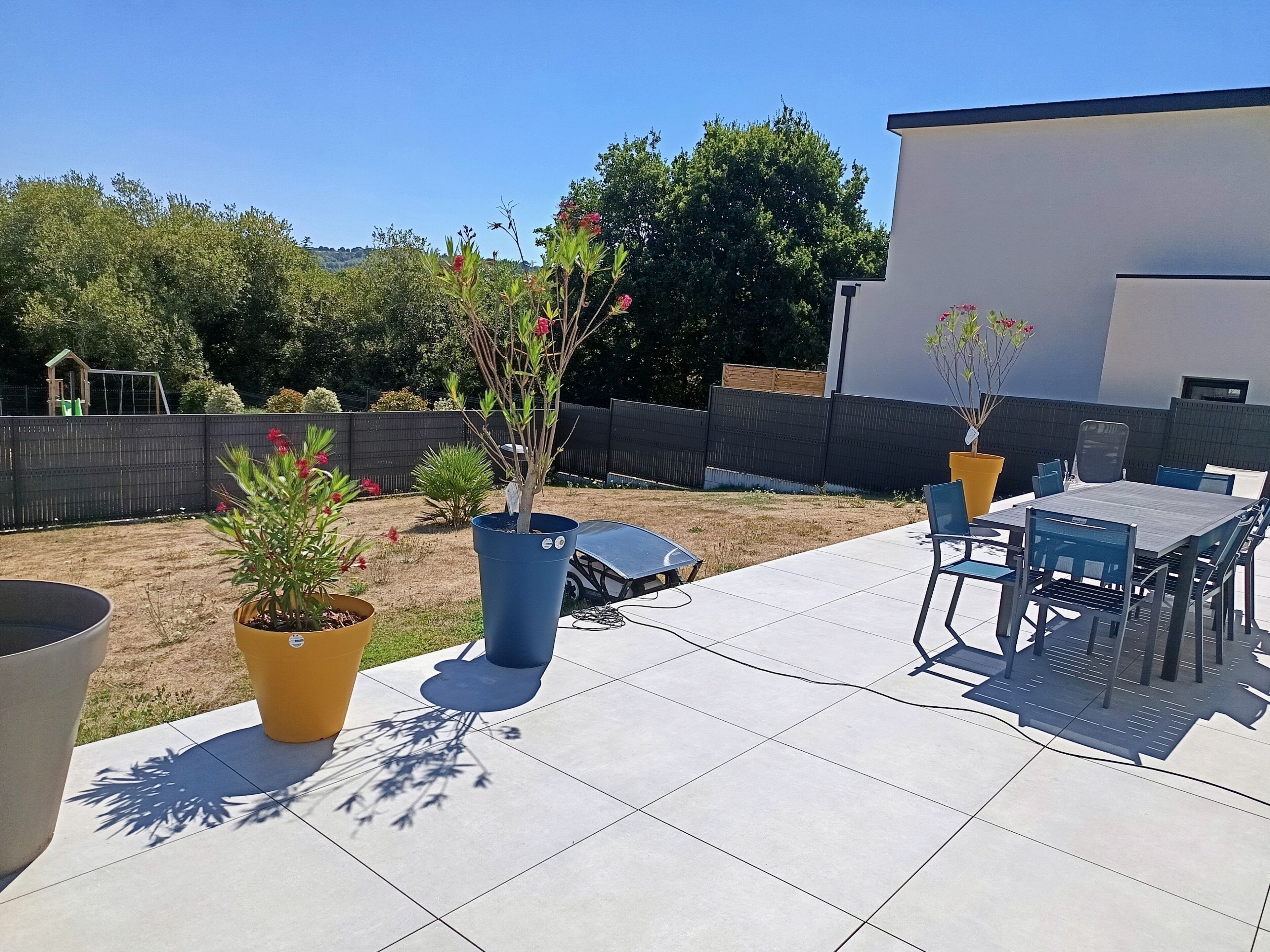 Een terras buiten met een tafel en stoelen, plantenpotten en een omheind erf.