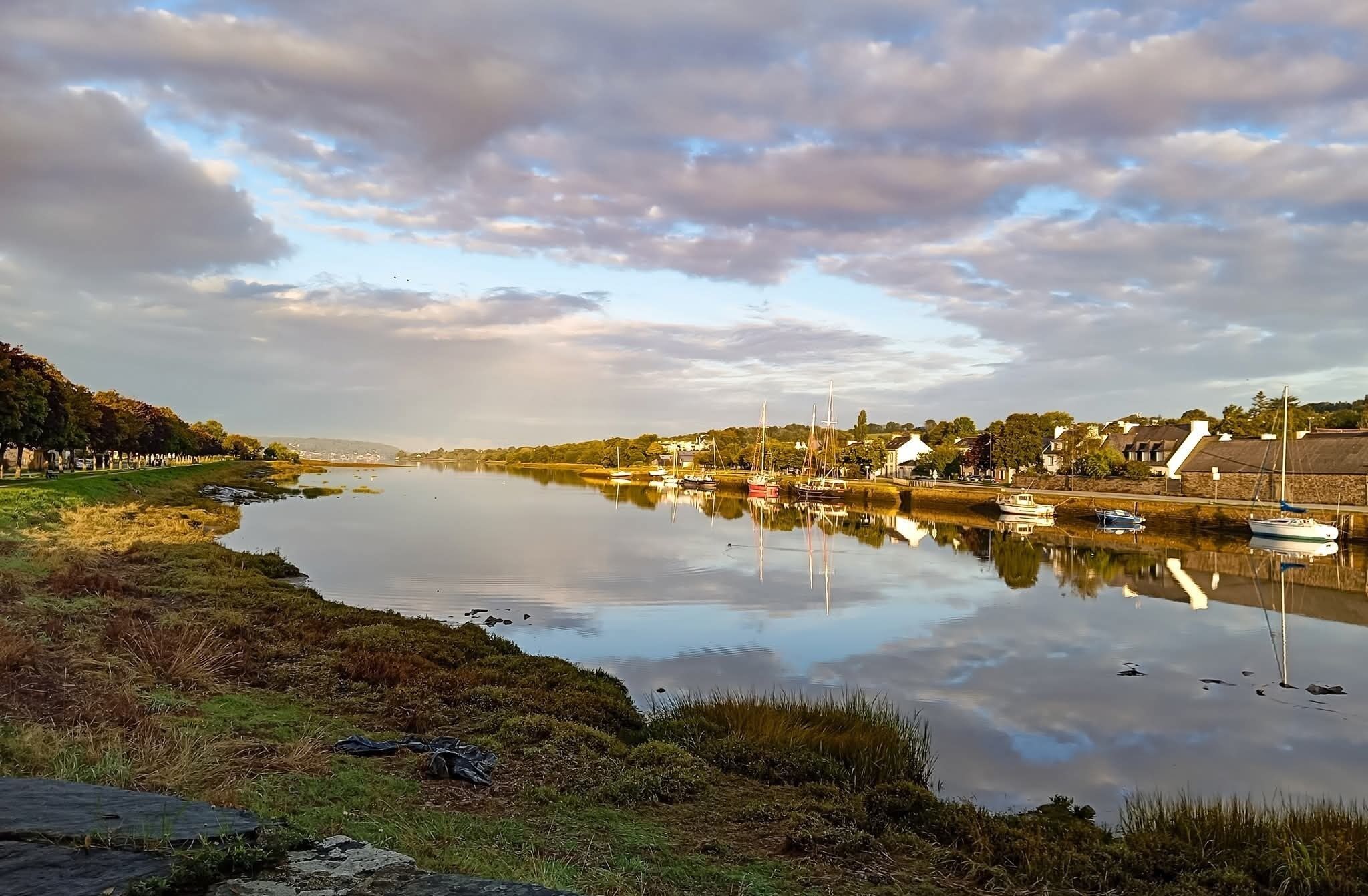 A calm river with boats, a grassy bank, and buildings in the background.