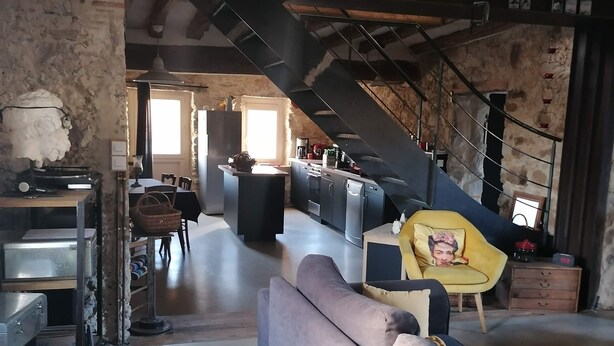 A modern kitchen with a central island, a dining area with a sofa, and a staircase leading to an upper level. - Maison Ancienne au Calme (Soulaire-et-Bourg, France)