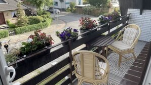 Un balcón con sillas de mimbre y una mesa, con vista a una calle con casas y una carretera.