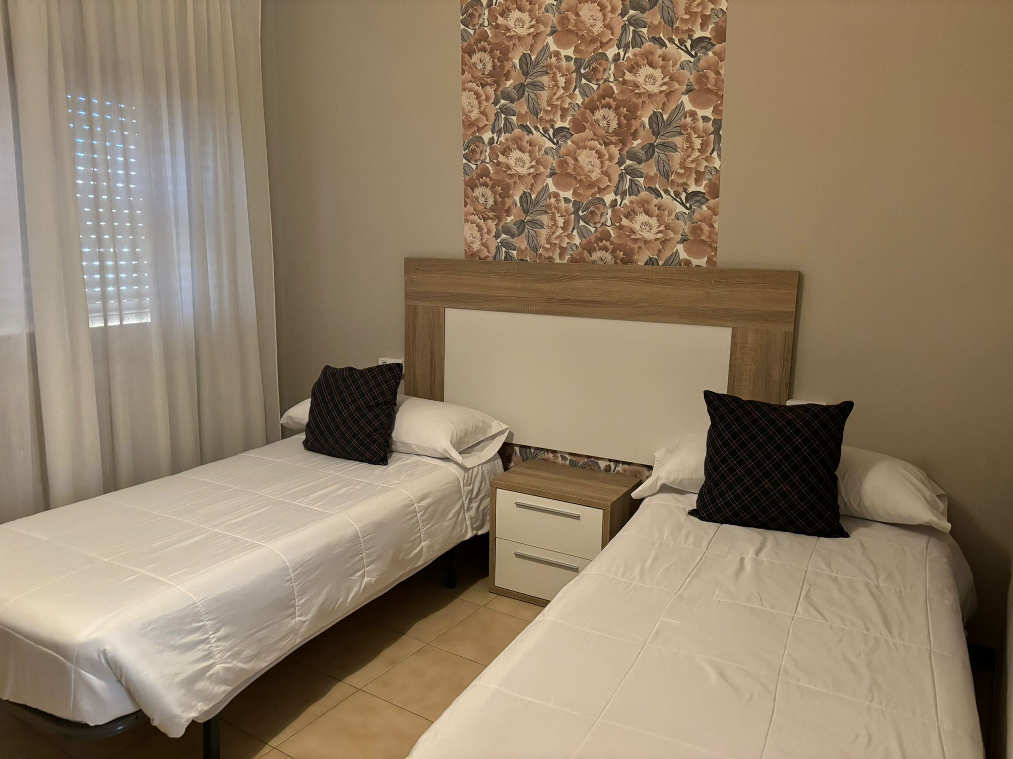 Une chambre d’hôtel avec deux lits simples, une table de chevet, une tête de lit en bois et un papier peint à motifs floraux.