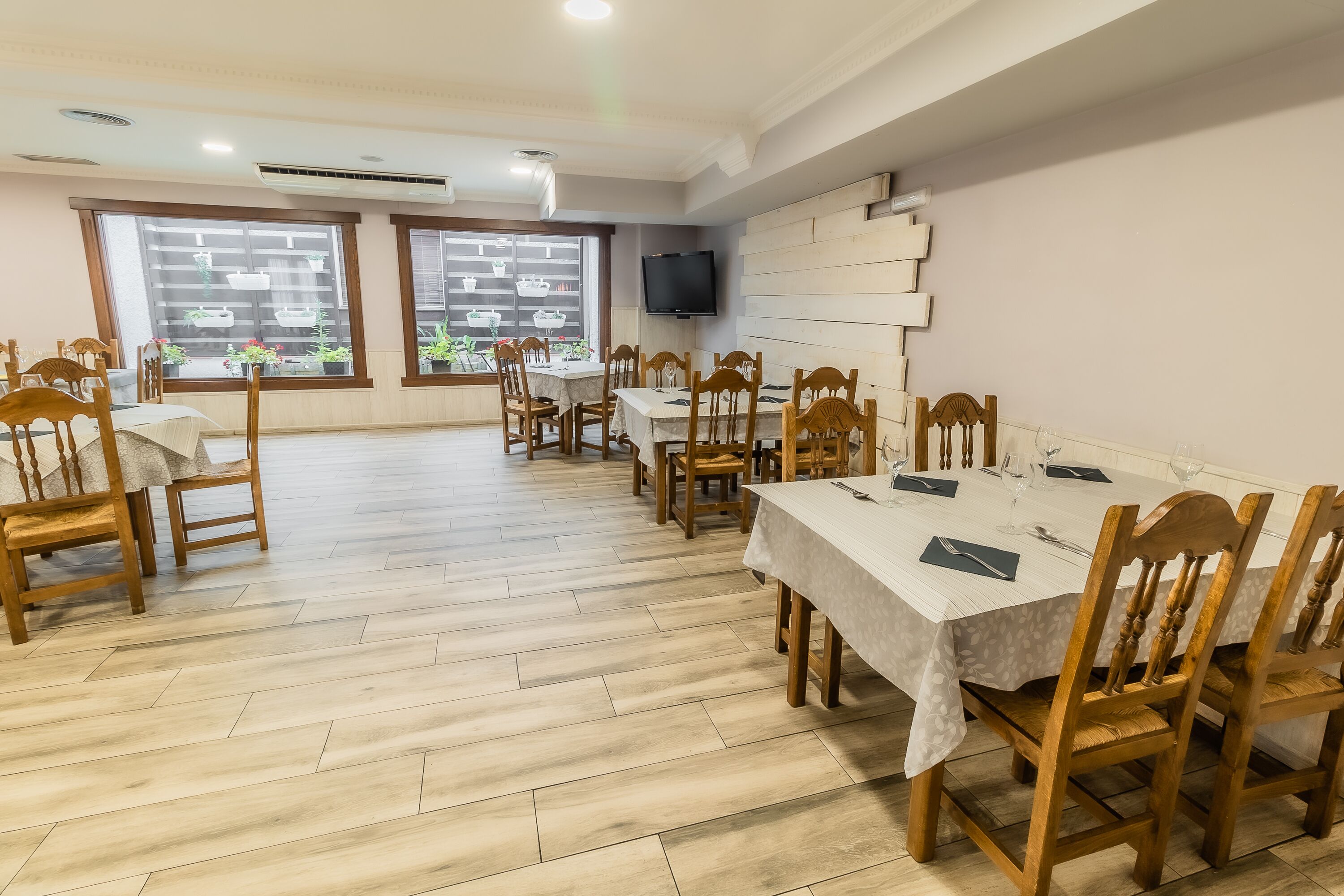A dining area with wooden chairs and tables, a flat-screen TV on the wall, and large windows with potted plants.
