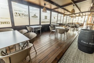 A restaurant interior with wooden flooring, white tables and chairs, hanging pendant lights, and a black barrel labeled 'FINCA ANT'.