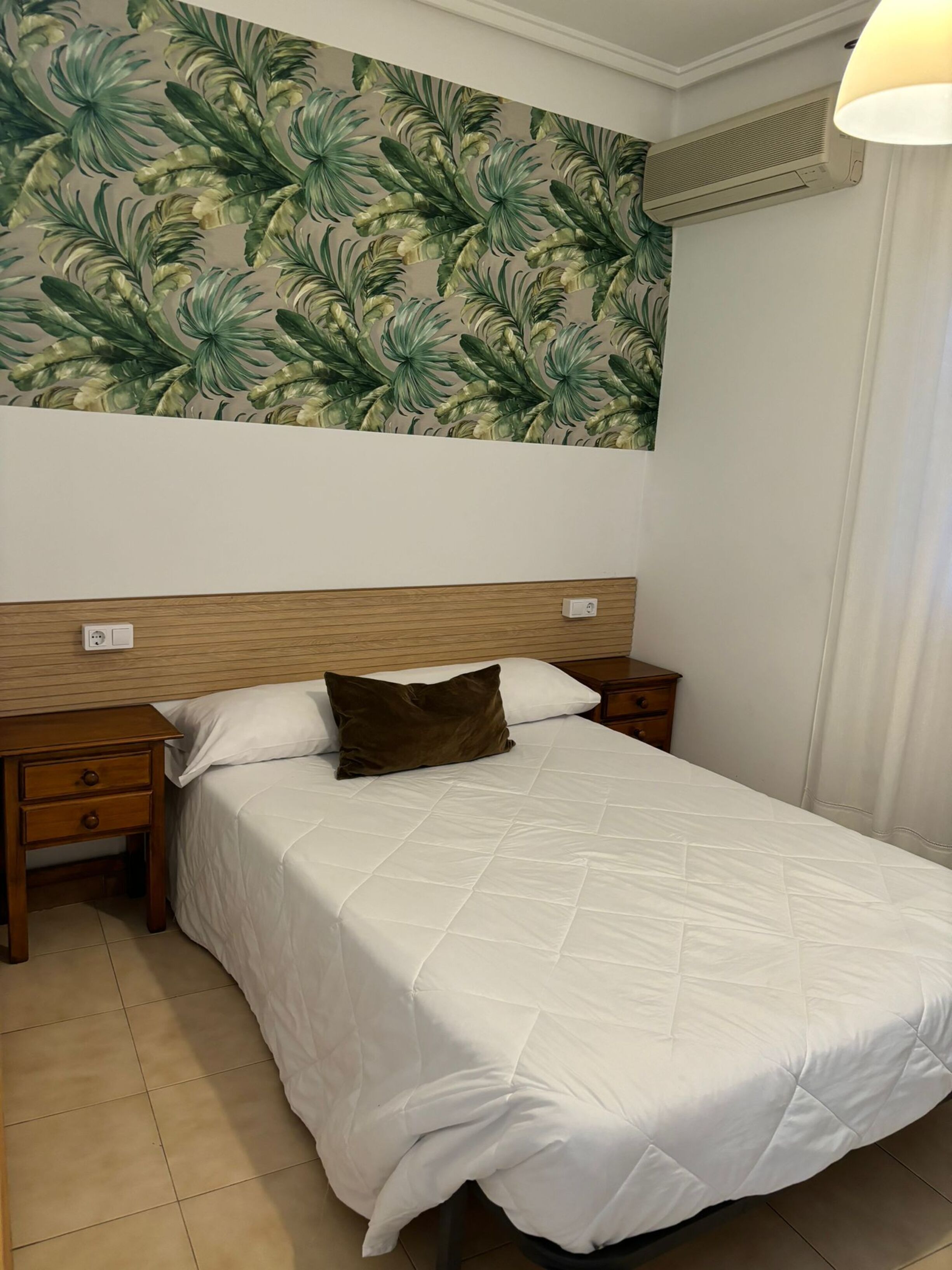 A hotel room with a bed, wooden headboard, white bedding, a nightstand, a drawer, and a wall with a tropical leaf pattern.