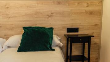 Quarto de hotel com cama, manta verde, cabeceira de madeira, mesa pequena e parede com mural de plantas.